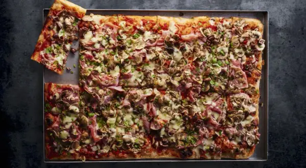 Pyszna pizza domowa przepis na blachę z szynką, pieczarkami i oliwkami, posypana serem i natką pietruszki.