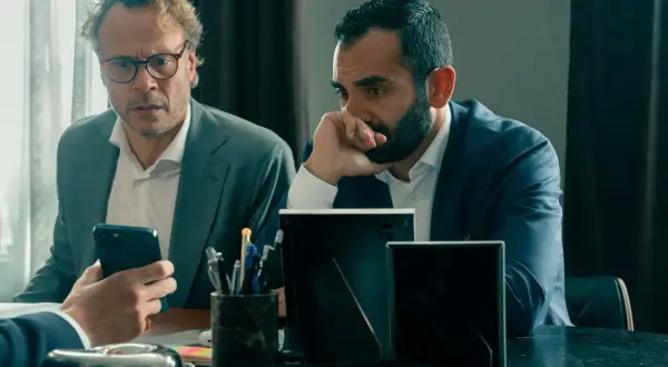 Två män i kostym sitter vid ett bord. Den ena mannen med glasögon tittar på sin mobiltelefon, den andra mannen funderar. Scen från "Hundarna" på SVT.