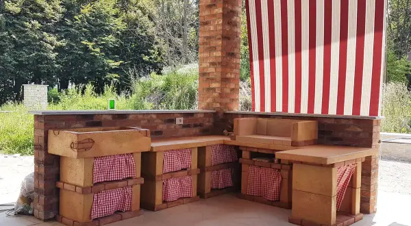 Cucina esterna in muratura fai da te con lavello, piano di lavoro e seduta. Tende a righe rosse e bianche proteggono lo spazio.