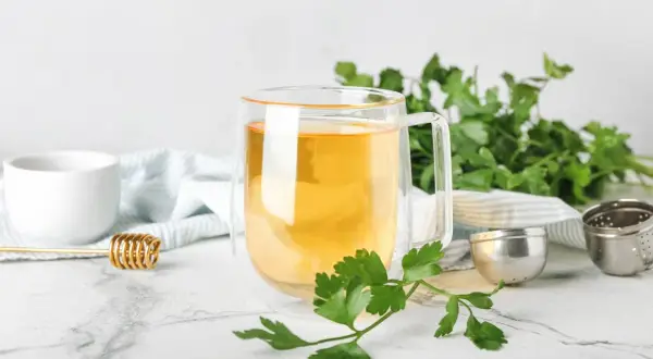 Aromatyczna herbata z pietruszki w szklance, obok świeże gałązki pietruszki, miód i zaparzacz.