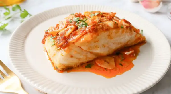 Delicioso bacalao a la llauna con salsa de tomate y ajo, servido en un plato blanco con un tenedor dorado.