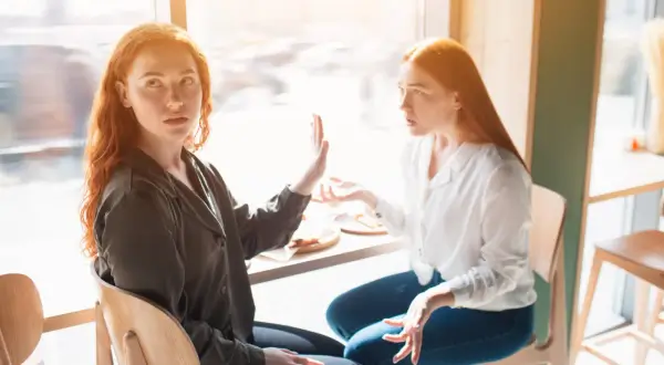 Zwei Frauen mit roten Haaren sitzen sich gegenüber. Eine hält die Hand hoch, als ob sie sagt: "Hör auf zu reden", wenn Kollegen nicht mit einem sprechen.