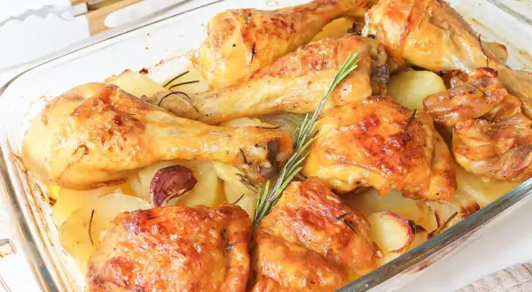 Muslos de pollo al horno con patatas, dorados y jugosos, acompañados de patatas asadas, ajo y romero en una fuente de cristal.