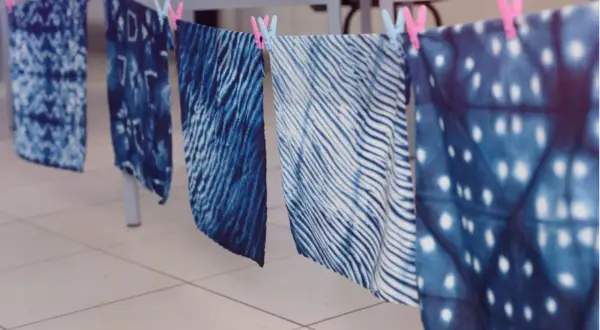 Stoffbahnen mit verschiedenen blauen Batik-Techniken trocknen an einer Leine. Muster wie Wellen, Punkte und abstrakte Formen sind sichtbar.