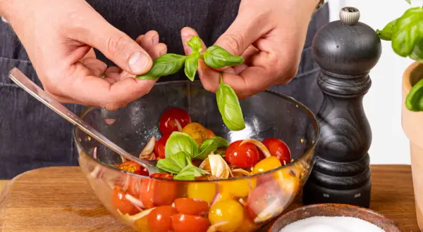 Hände geben Basilikumblätter in eine Schüssel mit Tomaten und Zwiebeln.