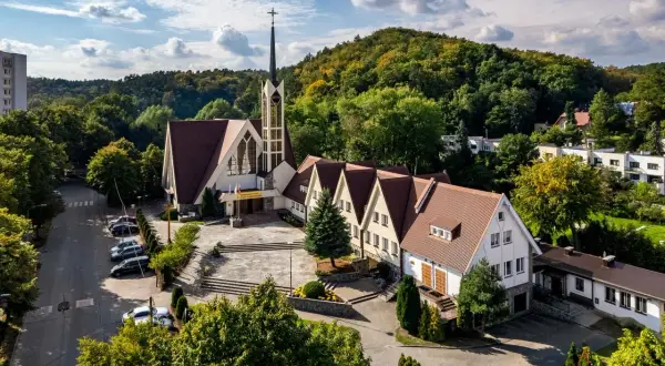 Nowoczesny kościół z wieżą i przylegającym budynkiem parafialnym, otoczony zielenią. W oddali widać wzgórza i zabudowania. To miejsce, gdzie historia spotyka się z teraźniejszością, jak w vii dwór.