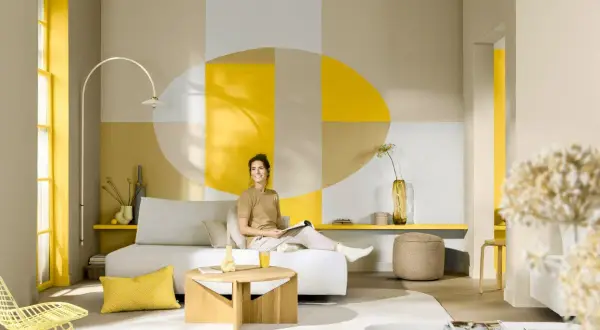 Eine Frau entspannt sich in einem hellen Wohnzimmer mit geometrischer Wandgestaltung. Welche Farbe passt zu Gelb? Hier: Beige, Grau und Weiß harmonieren perfekt.