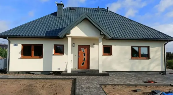 Nowoczesny dom kanadyjski z jasną elewacją, ciemnym dachem i drewnianymi oknami. Przed domem ścieżka z kostki brukowej i plac budowy.