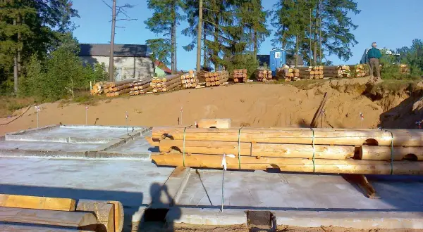 Budowa domu z bali: fundamenty, stosy drewna i pracownik na budowie.