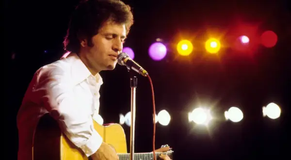 Artysta w białej koszuli gra na gitarze akustycznej, śpiewając do mikrofonu. W tle kolorowe światła sceniczne.