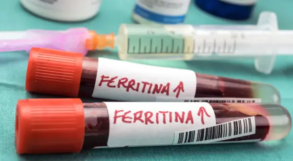 Dwie probówki z krwią oznaczone "Ferritina" i strzałką w górę, sugerujące potrzebę podniesienia ferrytyny. W tle strzykawka i fiolki.