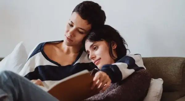Dwie kobiety przytulone na kanapie, jedna czyta, druga opiera głowę na jej ramieniu. W tle widać książki o lesbijkach.