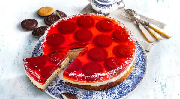 Pyszne ciasto z delicjami, galaretką i bitą śmietaną, ozdobione wiórkami kokosowymi. Obok leżą ciasteczka.