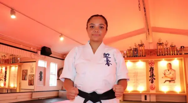 En kvinna med ett svart bälte i karate står redo i en dojo, omgiven av troféer och karaktärstecken.