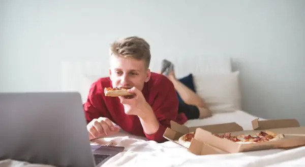 A young man eats pizza in bed while watching a laptop. Is it bad to go to sleep after eating? This scene suggests a late-night snack before bed.