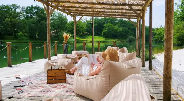 Détente sous le toit de chaume du whaka lodge, au bord d'un lac paisible. Une femme lit, entourée de poufs confortables.