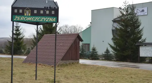 Tablica z napisem "Żeromszczyzna" i stylizowanym domkiem. Obok budynek z napisem "Centrum Edukacyjne Szklany Dom".