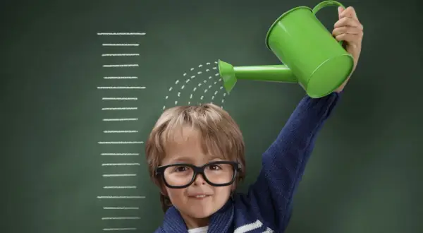 Niño con gafas riega su cabeza con una regadera verde, frente a una tabla de crecimiento que marca el percentil 10.