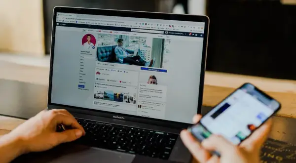 Osoba sprawdza na telefonie, jak sprawdzić logowania na FB, widząc profil na laptopie.