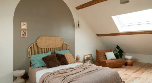 Een rustige, minimalistische slaapkamer met een rieten hoofdeinde, zachte plaids en een comfortabele fauteuil onder een dakraam.