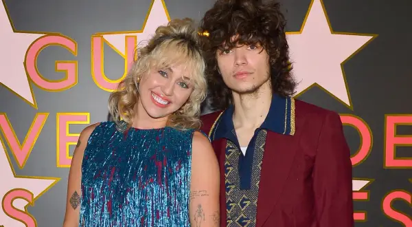 Miley Cyrus y Max Morando posan sonrientes. Ella lleva un vestido azul brillante con flecos, él un traje burdeos con detalles dorados.