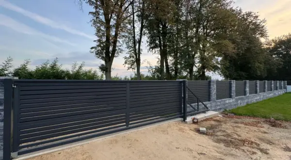 Nowe, nowoczesne ogrodzenie z ciemnoszarymi panelami i kamiennymi słupkami. W tle widać wysokie drzewa w linii ogrodzenia, tworzące naturalną barierę.
