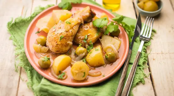 Tajín de pollo con patatas, aceitunas y limón, servido en plato rosa sobre mantel verde.