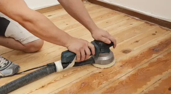 Un homme ponce un parquet en bois avec une ponceuse orbitale. C'est le bon moment pour le ponçage du parquet.