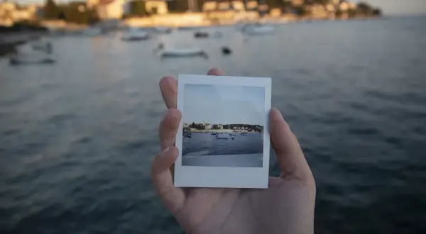 Dłoń trzyma zdjęcie jak z Instax z widokiem na zatokę z łodziami i miasteczkiem w tle.