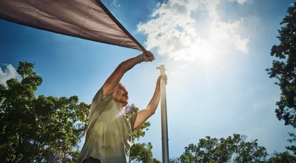 Man sätter upp ett solsegel mot en klarblå himmel med solens strålar som lyser.