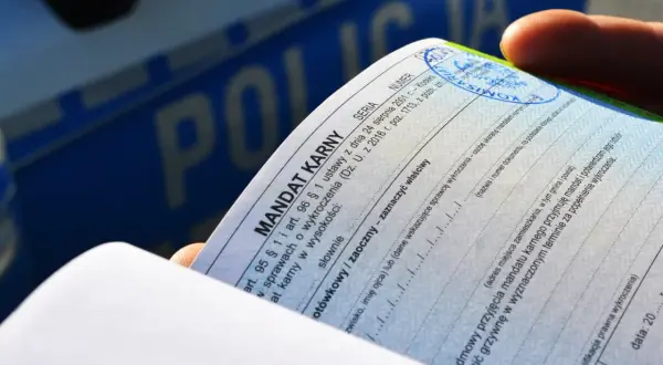 Dłoń trzyma mandat karny, dokument potwierdzający popełnienie wykroczenia. W tle widoczny napis "POLICJA".