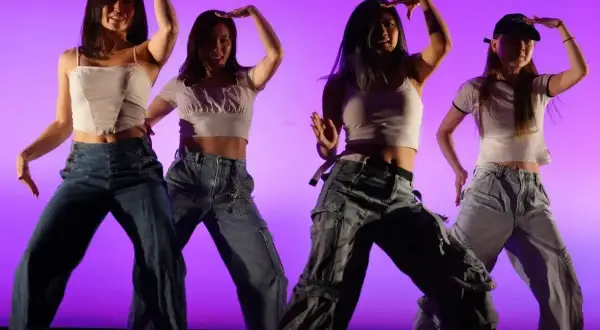 Cztery tancerki w luźnych jeansach i krótkich topach wykonują choreografię w stylu **key pop**.