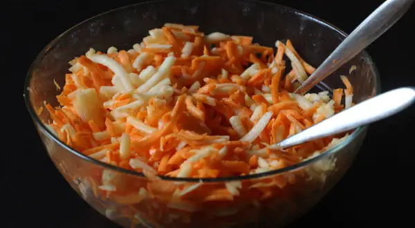 Schüssel mit geriebenen Karotten und Äpfeln, bereit für ein leckeres karottensalat rezept. Zwei Löffel warten darauf, den Salat zu servieren.