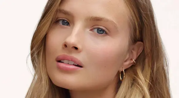 Visage d'une jeune femme aux yeux bleus et aux lèvres rosées, avec un maquillage subtil, style "makeup no makeup". Elle porte des boucles d'oreilles dorées.