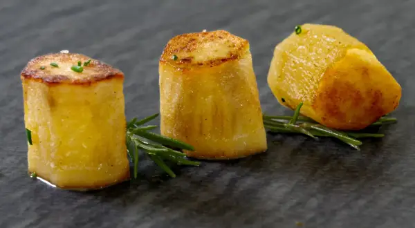 Drei goldbraune Fondantkartoffeln, garniert mit Rosmarin und Schnittlauch, auf dunklem Stein serviert.