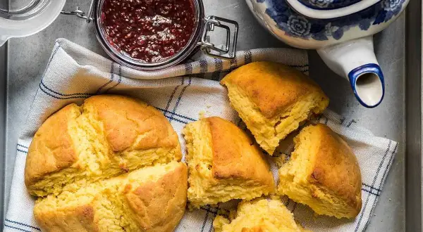 Gyllene scones majsmjöl, en burk hallonsylt och en blåvit tekanna. Perfekt för en mysig fika.