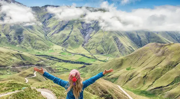 Mujer con brazos abiertos admira un paisaje montañoso verde, como si estuviera en la cima de la piedra del molino, con nubes y un camino sinuoso.
