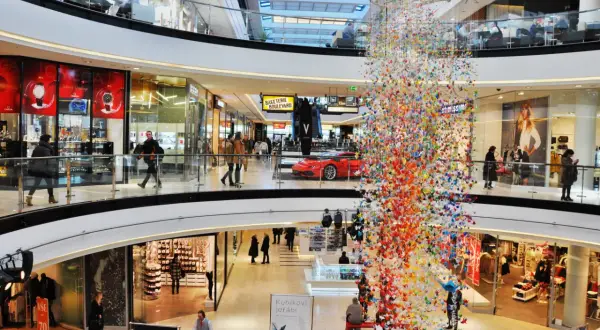 Bunte Papierkraniche schmücken ein Einkaufszentrum in Prag. Menschen bummeln durch die Gänge, vorbei an Geschäften und einer roten Sportwagen-Auslage.