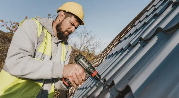 A roofer in a yellow hard hat and hi-vis vest uses a drill for metal roof repair.