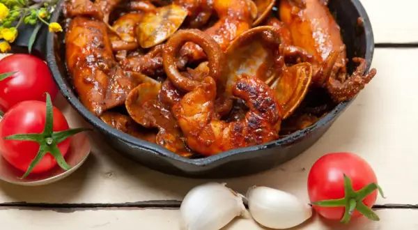 Un plat de calamars à l'armoricaine mijoté dans une sauce rouge, servi dans une poêle en fonte, avec des tomates et de l'ail à proximité.