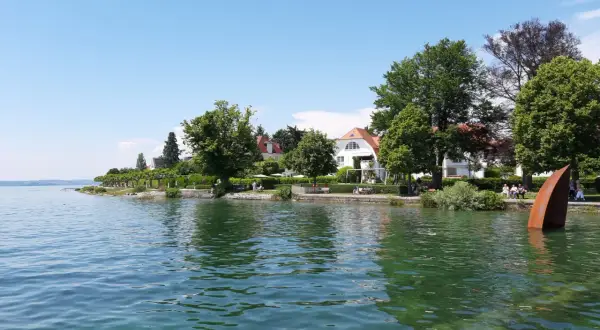 Die Hagnau Promenade am Bodensee mit weißen Häusern, grünen Bäumen und einer rostigen Skulptur im Wasser.