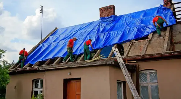 Pracownicy na dachu, przykrytym niebieską plandeką, wykonują prace budowlane. Może to być próba ukrycia **zmiany konstrukcji dachu bez pozwolenia**.