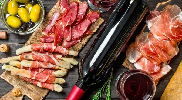 Un bicchiere di vino rosso, una bottiglia, salumi, olive e grissini avvolti nel prosciutto, un'ambientazione perfetta per la vinificazione in rosso.