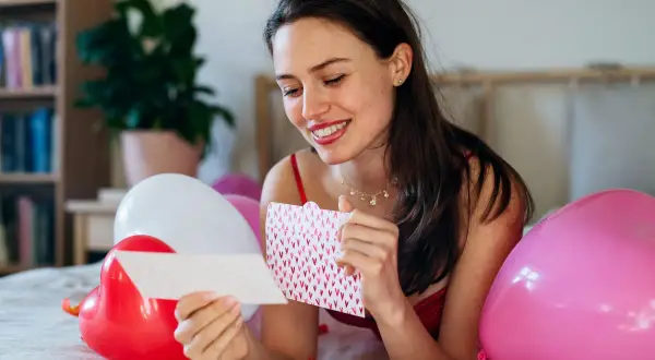 Uśmiechnięta kobieta czyta kartkę z serduszkami, otoczona balonami. Może to być inspiracja na wiersze dla ukochanego mężczyzny.