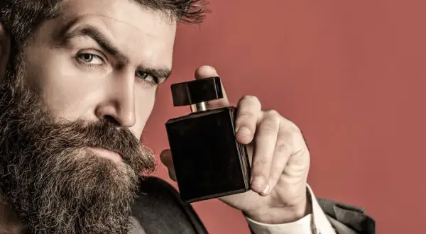 Mężczyzna z brodą trzyma czarną buteleczkę perfum na czerwonym tle.