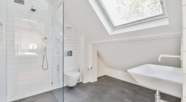 Moderno baño en buhardilla con ducha de cristal, inodoro y lavabo bajo una claraboya.