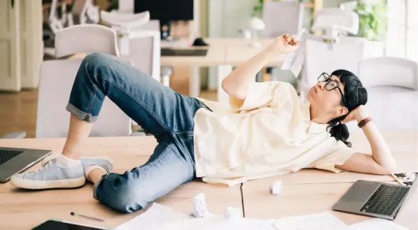 En person ligger utsträckt på ett skrivbord, omgiven av papper och laptops. Klädd i jeans och en ljus skjorta, ser hen ut att fundera över en papperslapp, en bild av en dysfunktionell arbetsplats.