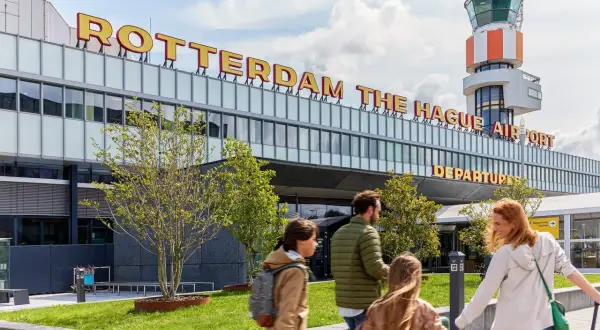 Rodzina zmierza na lotnisko Rotterdam The Hague Airport. W tle widoczna wieża kontroli lotów i napis z nazwą lotniska.