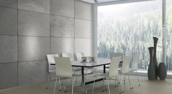 Esszimmer mit moderner Beton Optik Wandverkleidung, weißem Tisch und Stühlen, sowie einem Panoramafenster mit Blick auf einen Wald.
