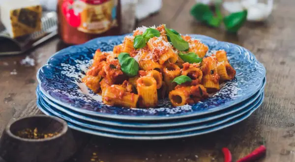 En tallrik med rigatoni all'arrabiata, toppad med basilika och parmesan. Bredvid syns en burk Mutti-tomatsås, vitlök och chilifrukter.
